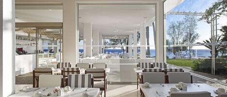 Ein modernes Restaurant mit Meerblick, ausgestattet mit gestreiften Stühlen und Tischen, und einer Glaswand, die den Essbereich von der Küche trennt.