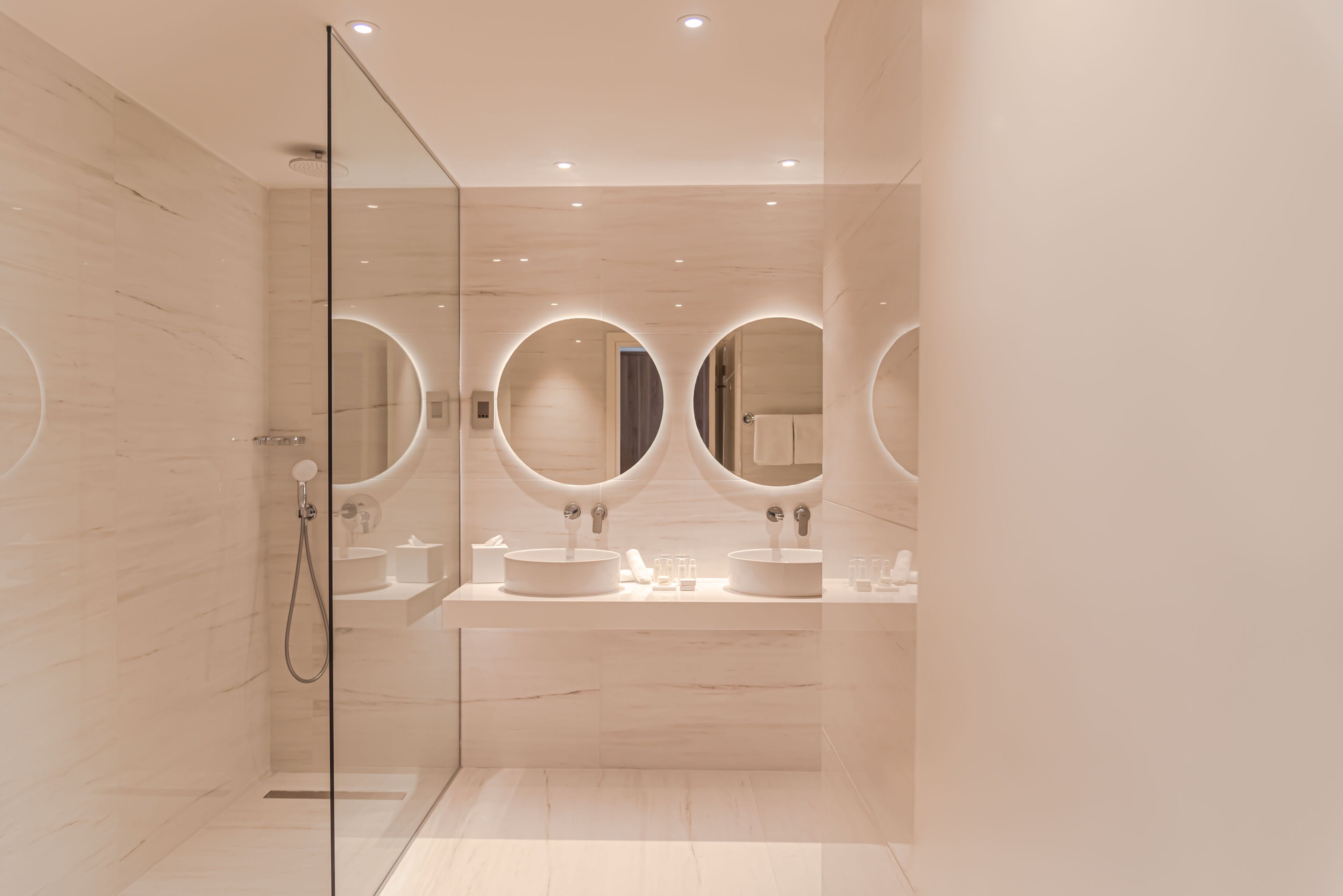 Een moderne badkamer met een glazen douchecabine, twee wastafels met ronde spiegels en een marmeren vloer.