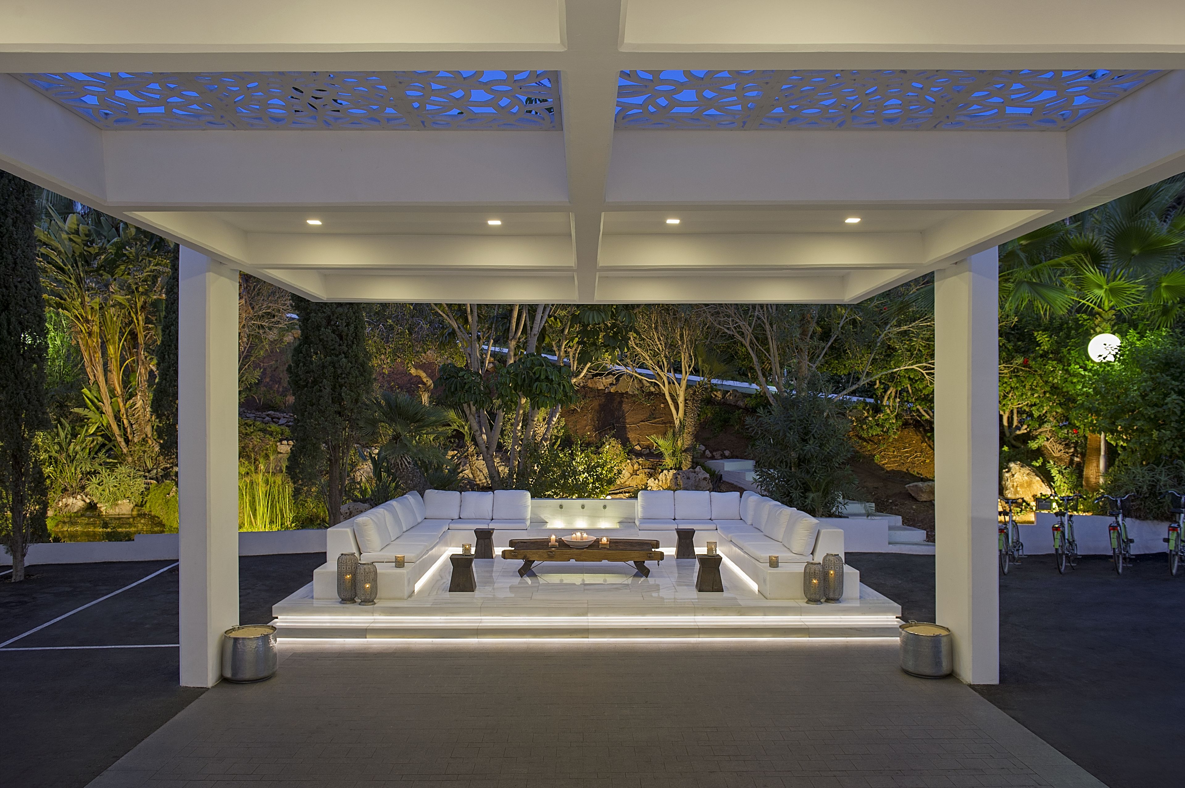 Un espace extérieur aménagé avec un canapé blanc et une table basse, entouré de plantes et d’arbres.