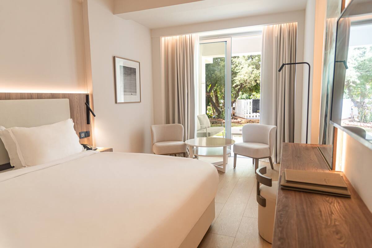 A hotel room with a large bed, a desk, two chairs, a small table, and a view of the outdoors.