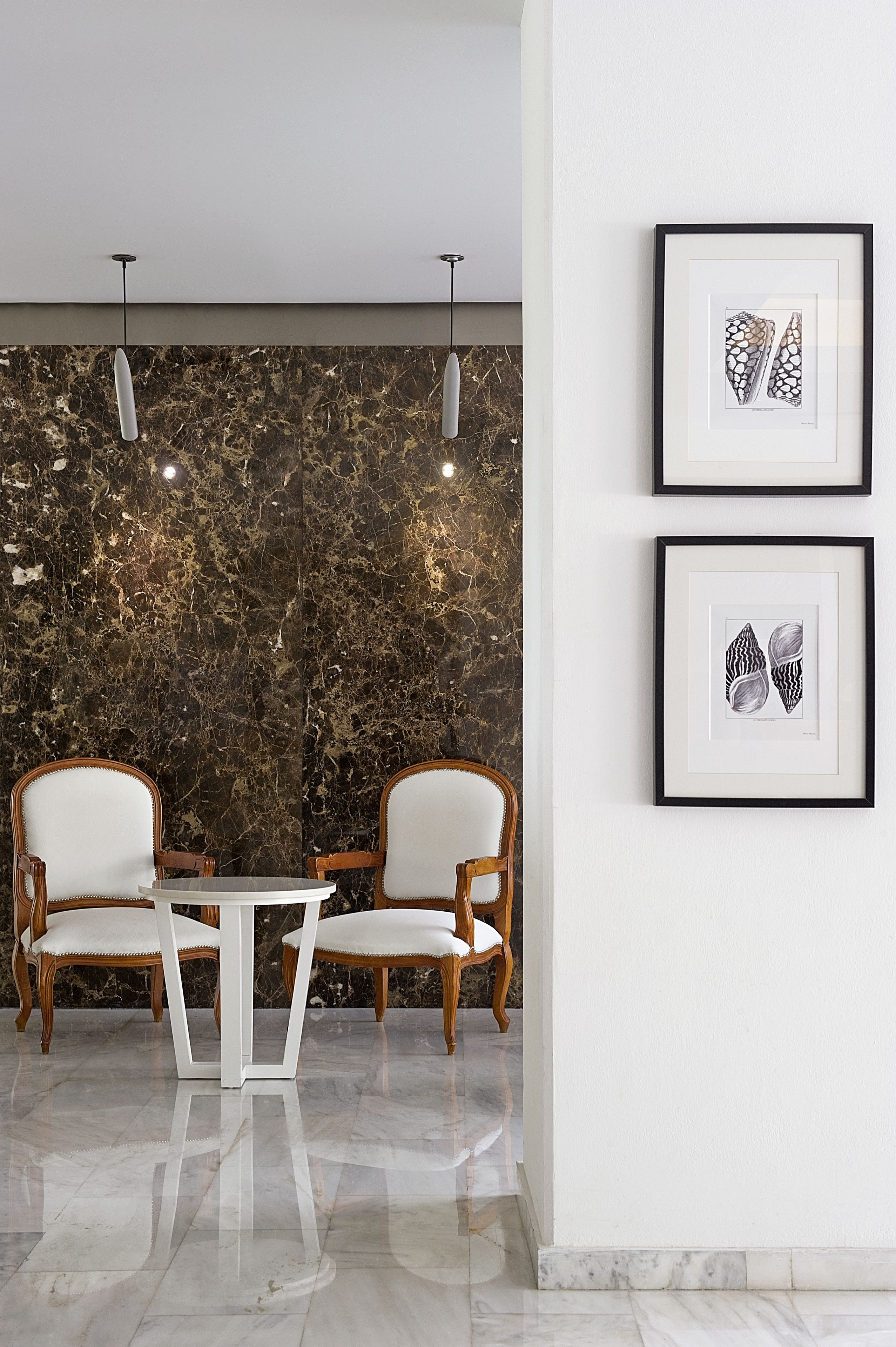 A dining area with a marble wall, two wooden chairs, a white table, and framed artwork on the wall.