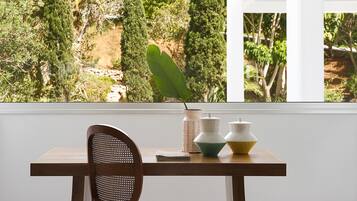 A wooden table with a woven chair, a potted plant, and decorative vases by a window overlooking a garden.
