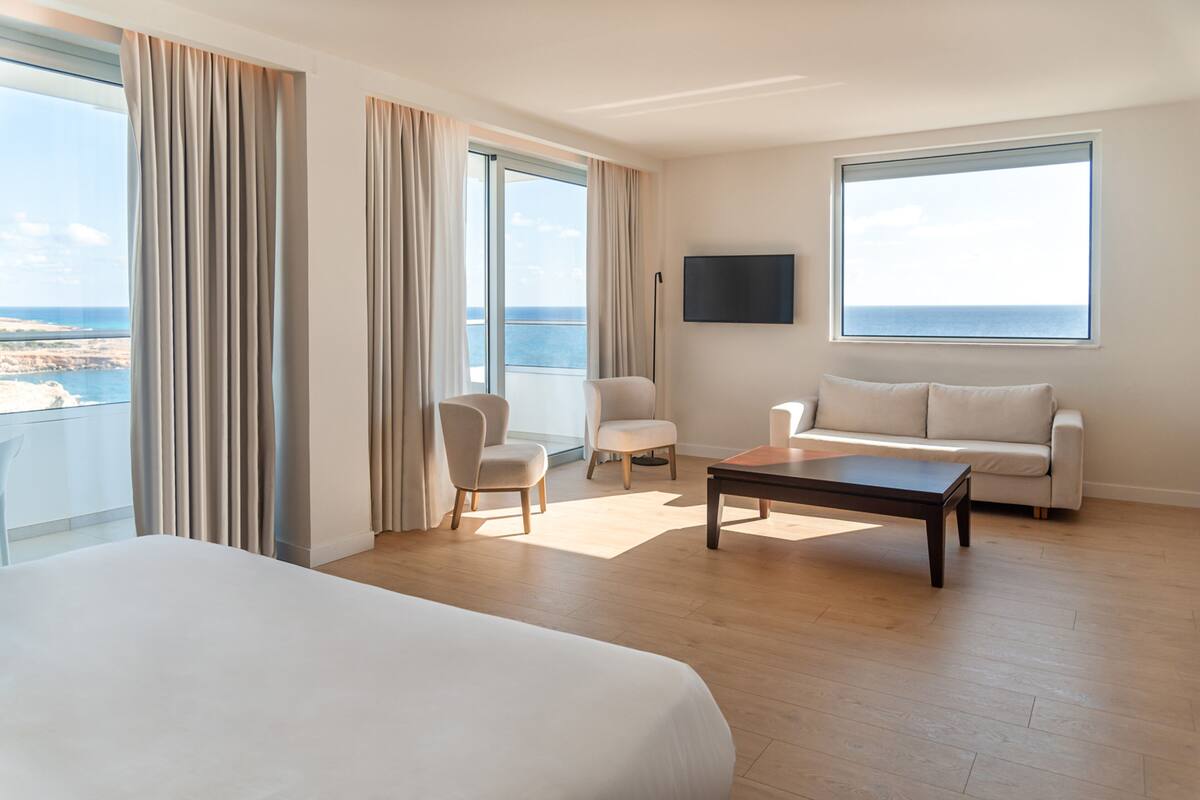 A hotel room with a large bed, a sofa, a coffee table, and a TV. There is a balcony with a view of the sea.