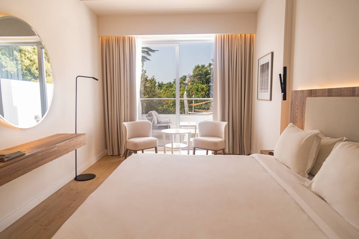 A hotel room with a large bed, a round mirror, a small table with two chairs, and a balcony with curtains.
