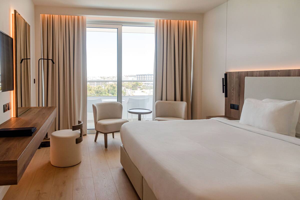 A modern hotel room with a large bed, two armchairs, a small round table, and a TV on a wooden console.
