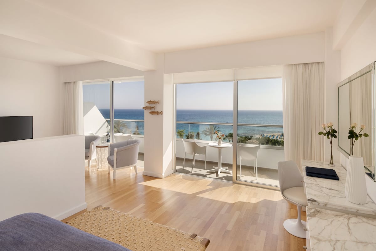 A modern hotel room with a large balcony, a TV, a seating area, and a view of the sea.