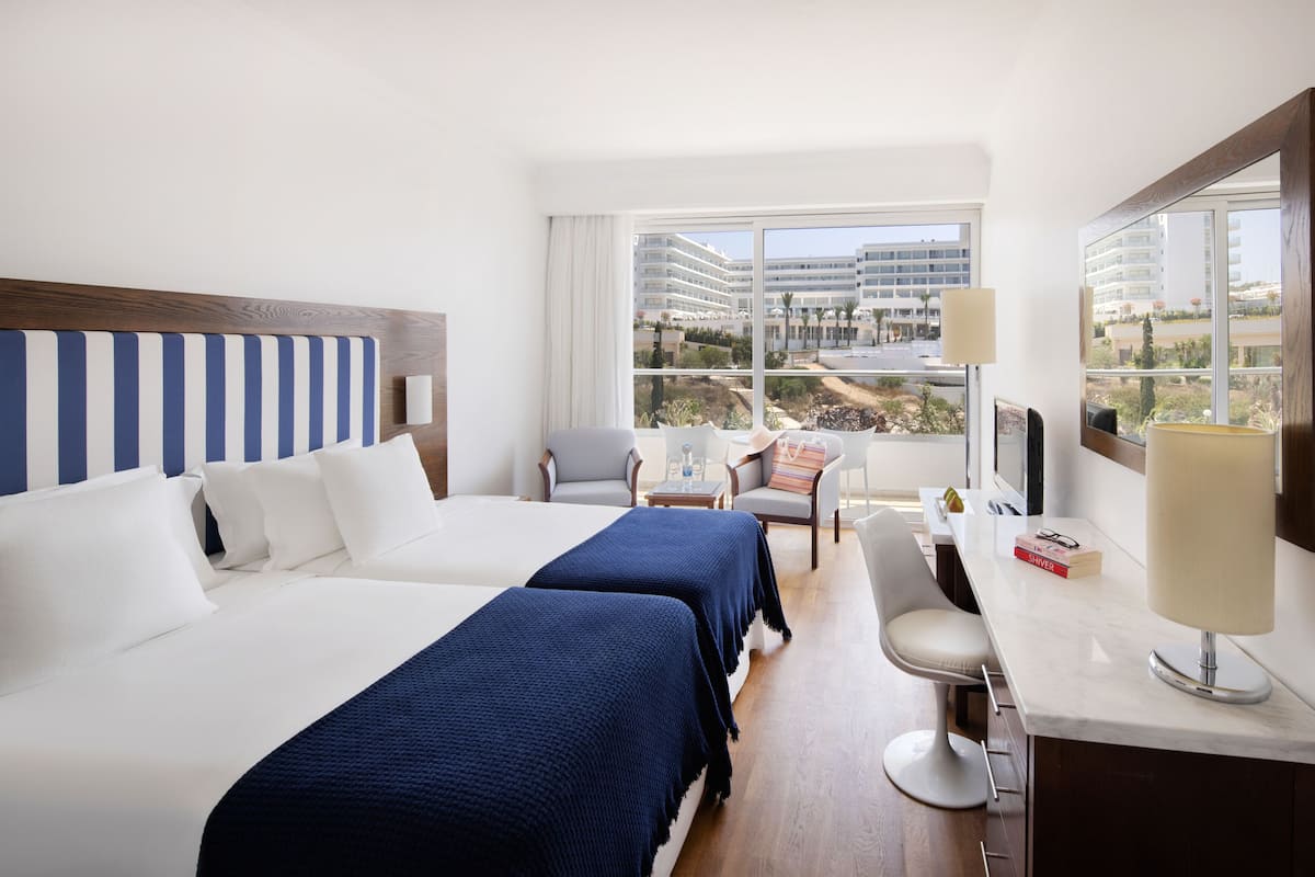 A hotel room with two beds, a sitting area with a chair and a table, a lamp, and a view of a building through large windows.
