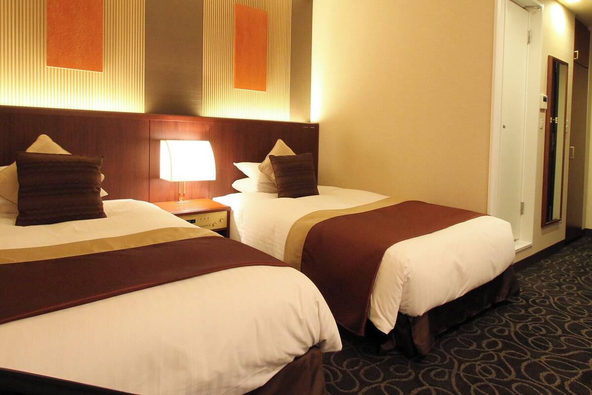 Dos camas de hotel juntas, con cabeceros, mesitas de noche con lámparas y suelo alfombrado.