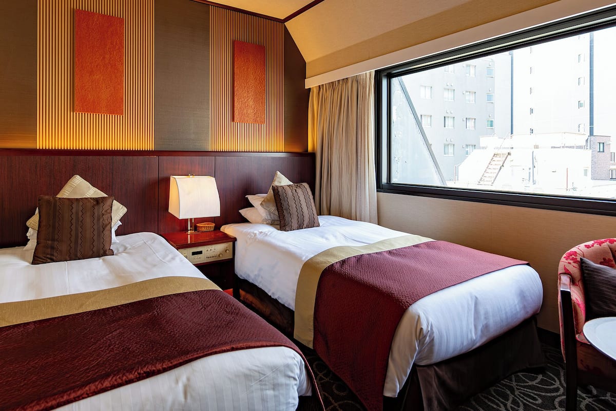 Habitación de hotel con dos camas, una ventana con vista a la ciudad y una mesita con una silla.