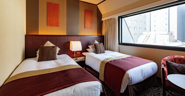 A hotel room with two beds, a window with a city view, and a small table with a chair.