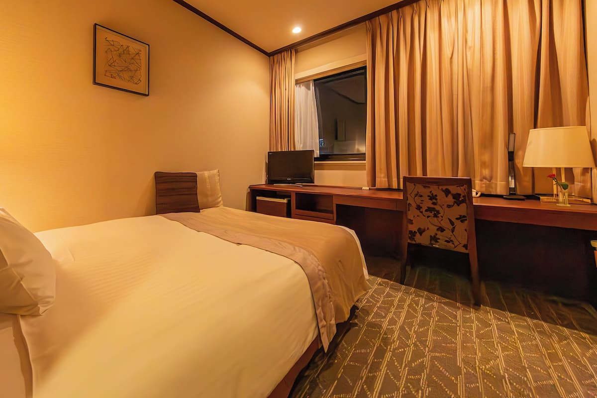 Una habitación de hotel con cama, escritorio, silla, lámpara y ventana con cortinas.
