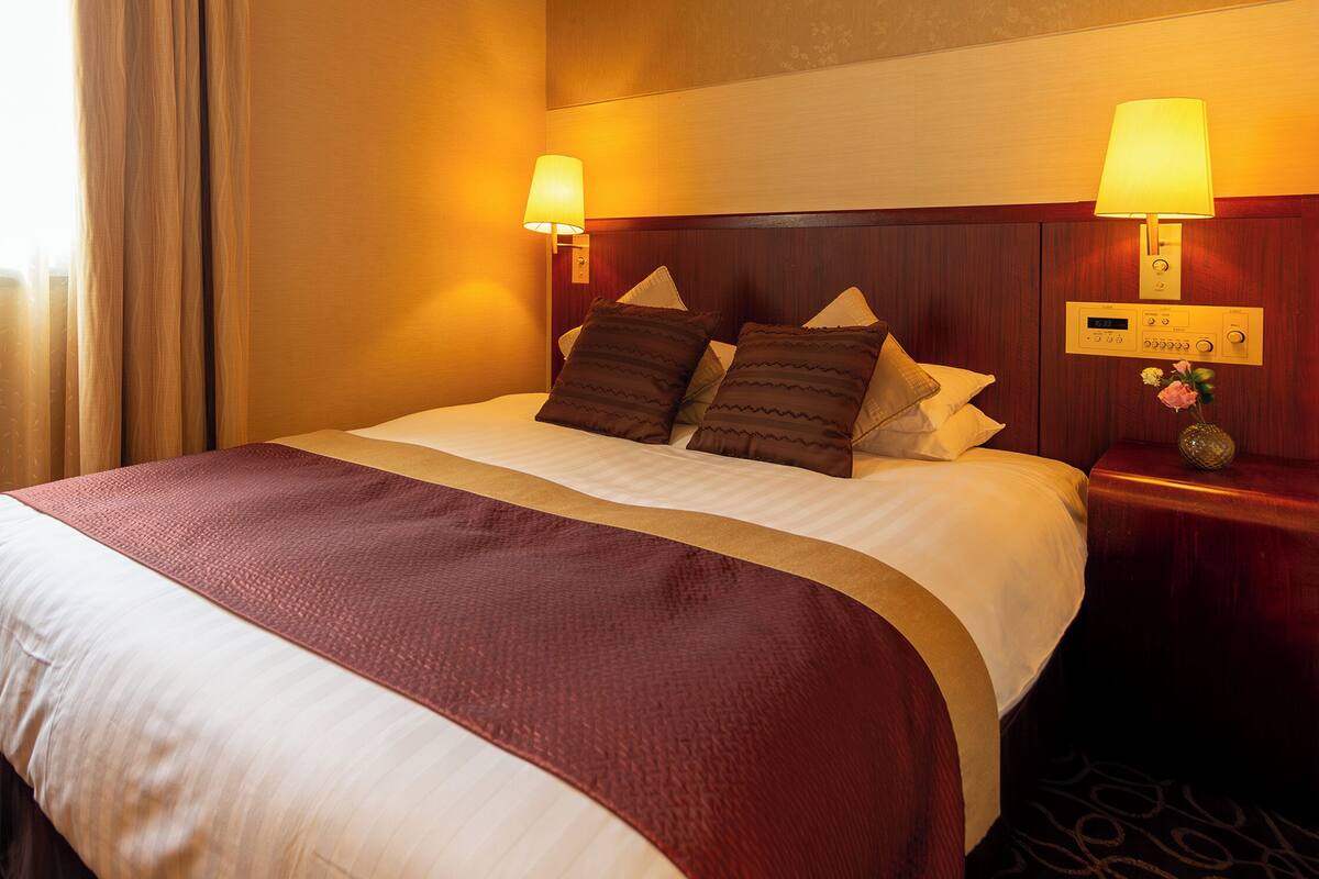 Habitación de hotel con una cama grande, dos lámparas de noche, un cabecero de madera y un sillón tapizado con motivos florales.