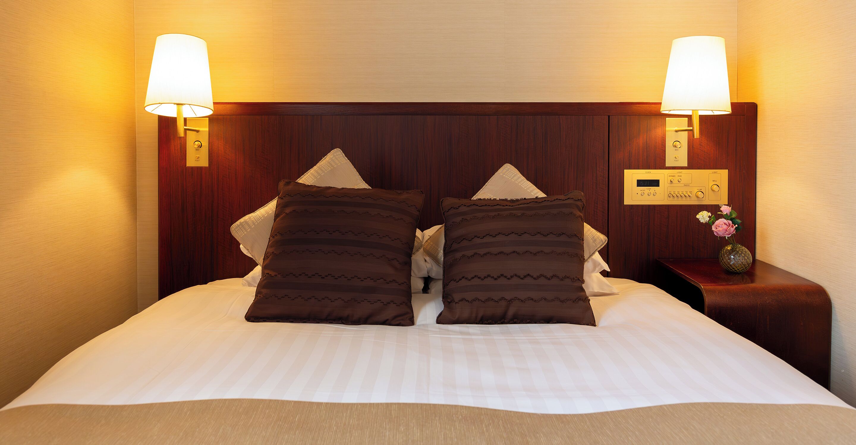 A neatly made bed with two decorative pillows, a bedside table with a clock, and wall-mounted lamps.