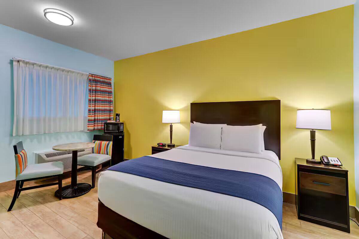 A hotel room with a large bed, two bedside lamps, a small table with a chair, a TV, and a window with curtains.