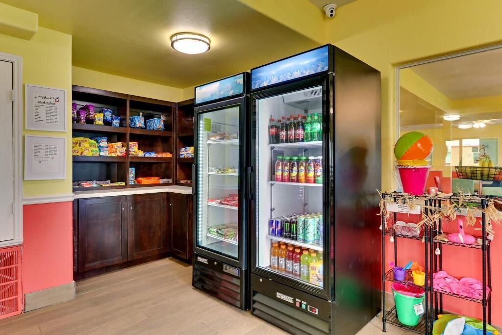 A room with a refrigerator stocked with beverages, a shelf with snacks, and a rack with cleaning supplies.