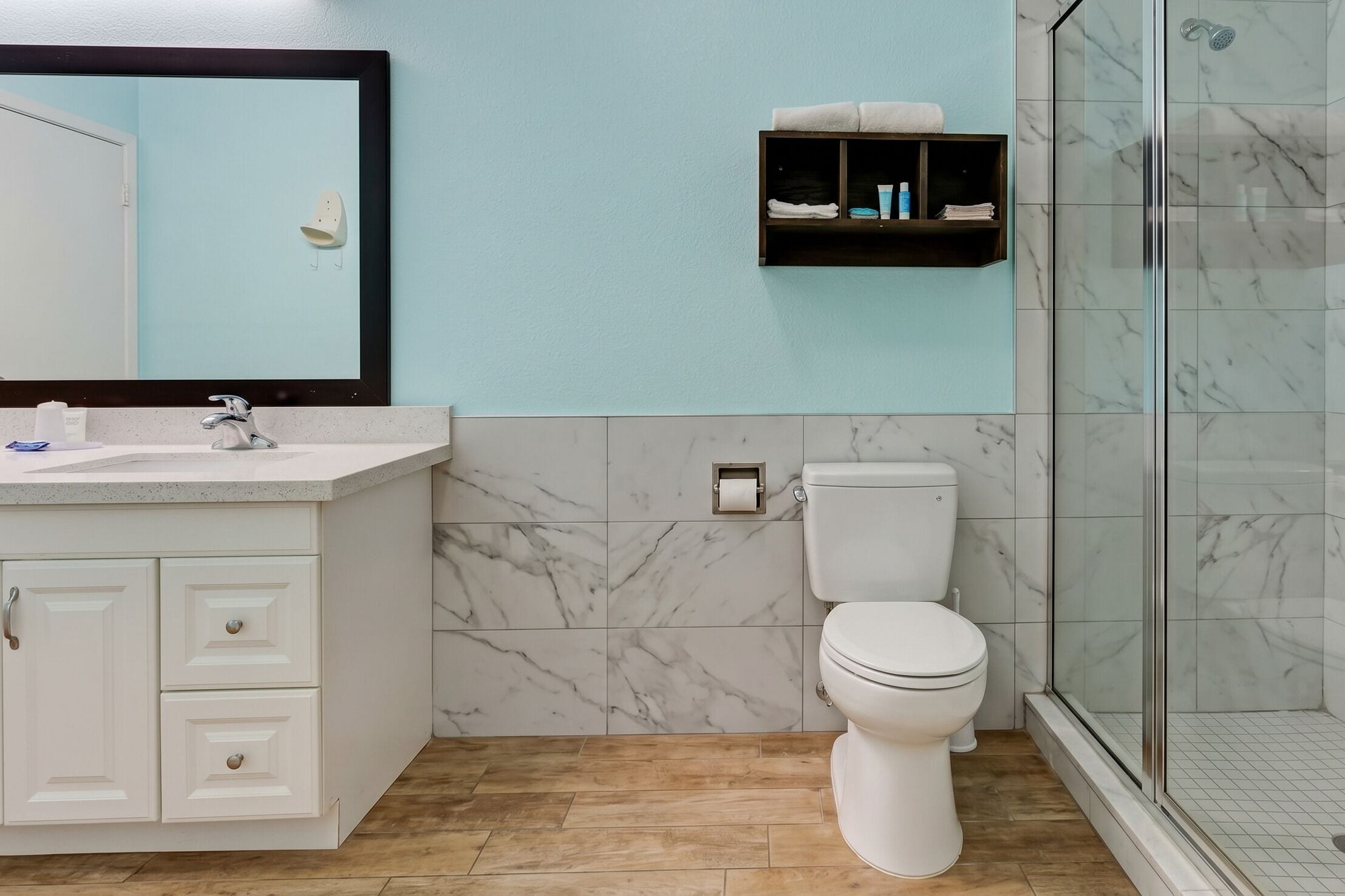 Baño con lavamanos blanco, baldosas de mármol, inodoro, cabina de ducha con mampara de vidrio y estante para toallas.