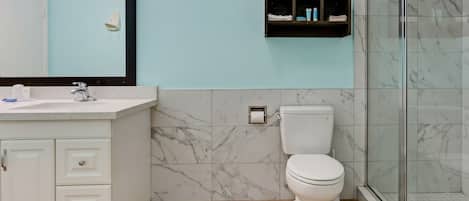 A bathroom with a white sink, marble tiles, a toilet, a glass shower enclosure, and a towel shelf.