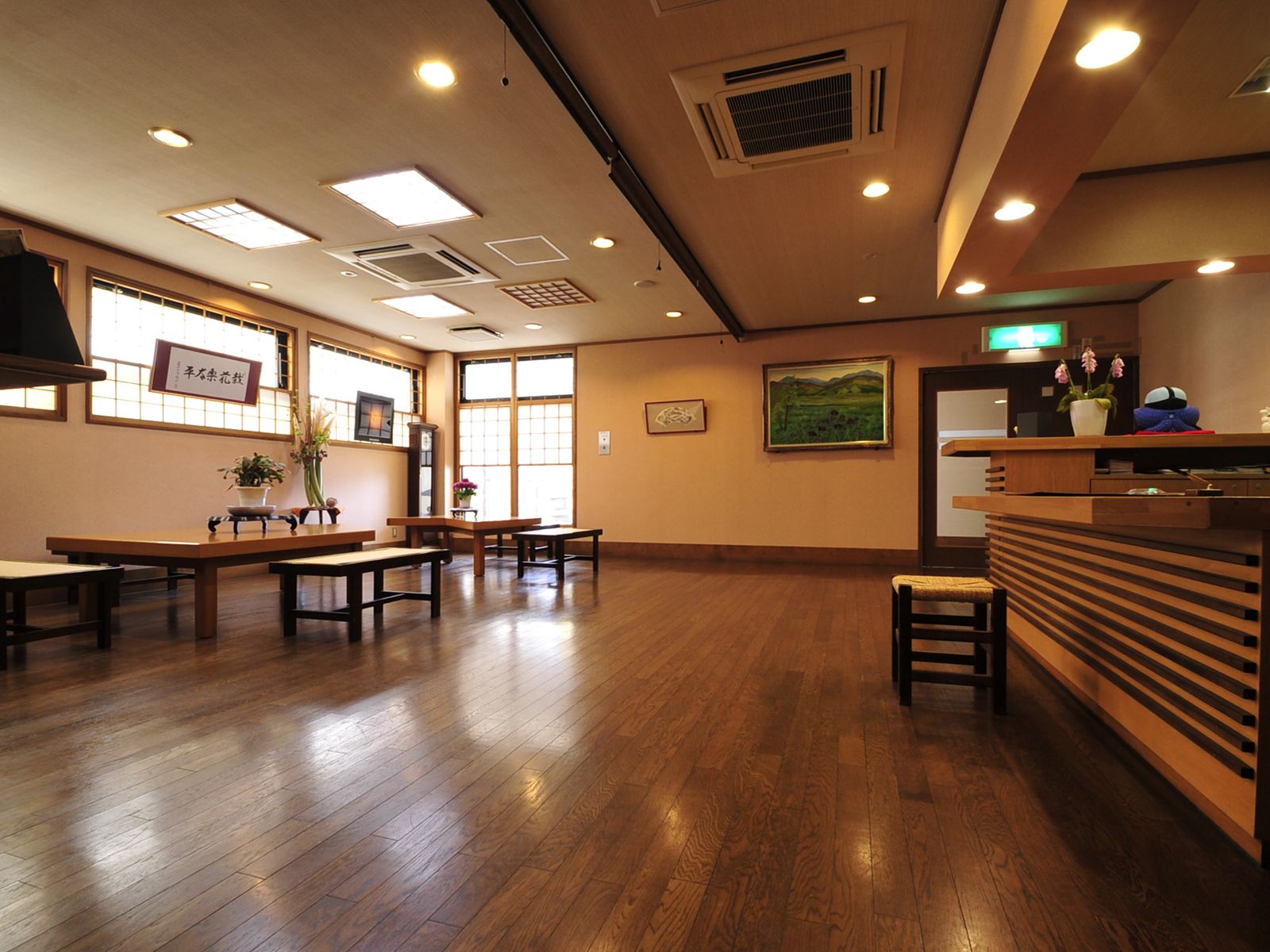 A spacious room with wooden flooring, multiple tables and benches, and a reception desk with a plant.