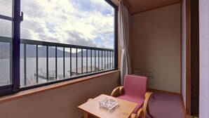 A small room with a window overlooking a body of water and mountains. There is a wooden table with a glass container on it, a pink chair, and a wooden bench. - Nikko Sansui (Nikko)