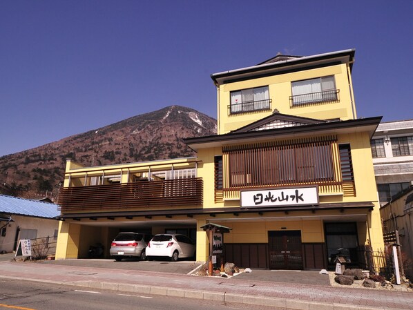 A two-story building with a yellow facade and a sign that reads "Yamato Mountain". There are two cars parked in front, and a mountain is visible in the background. - Nikko Sansui (Nikko)