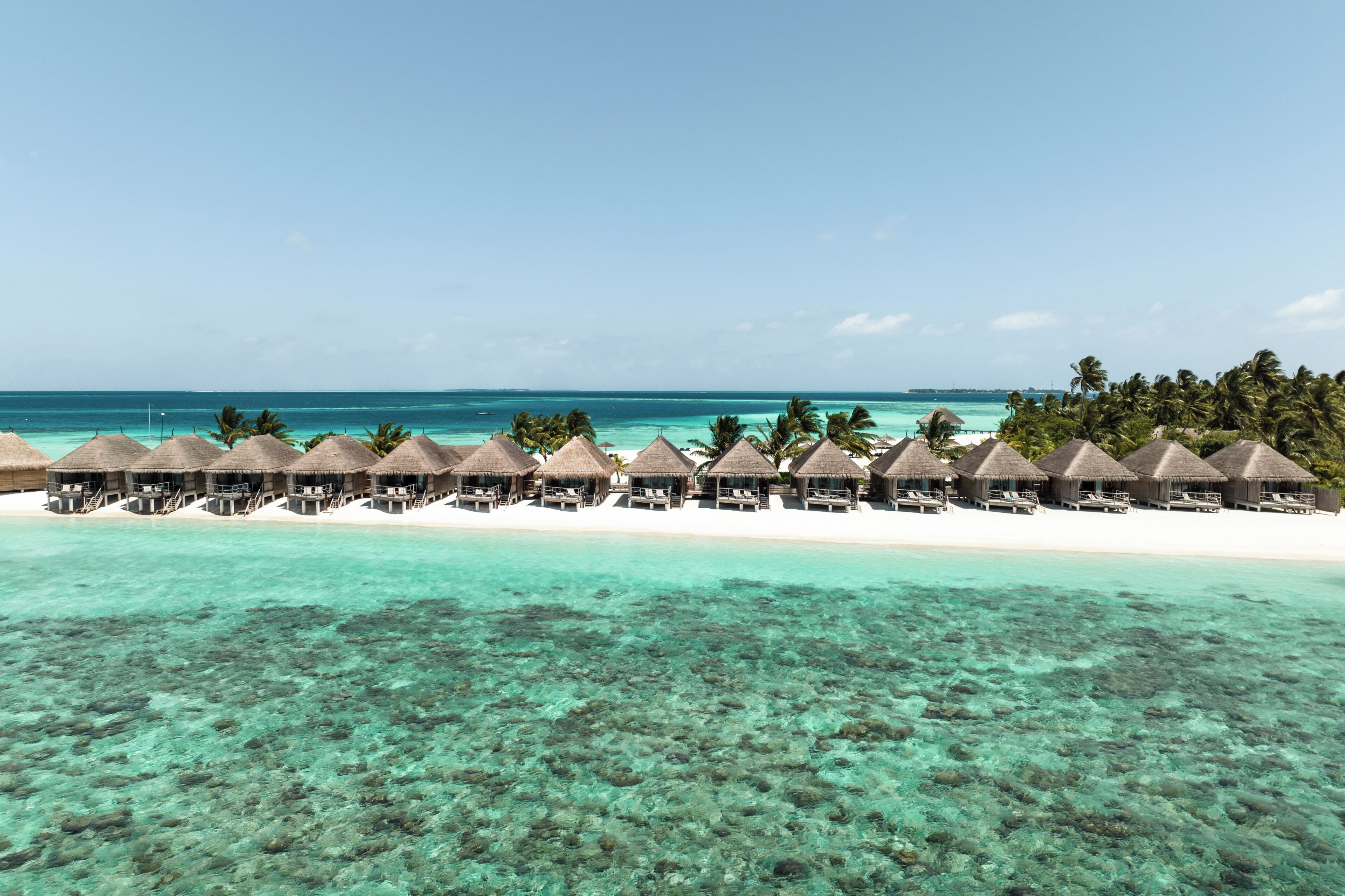 Ett lyxigt resorterbjudande med bungalows som ligger över vattnet, med tak av halmtak, kristallklart turkost vatten och en vit sandstrand.