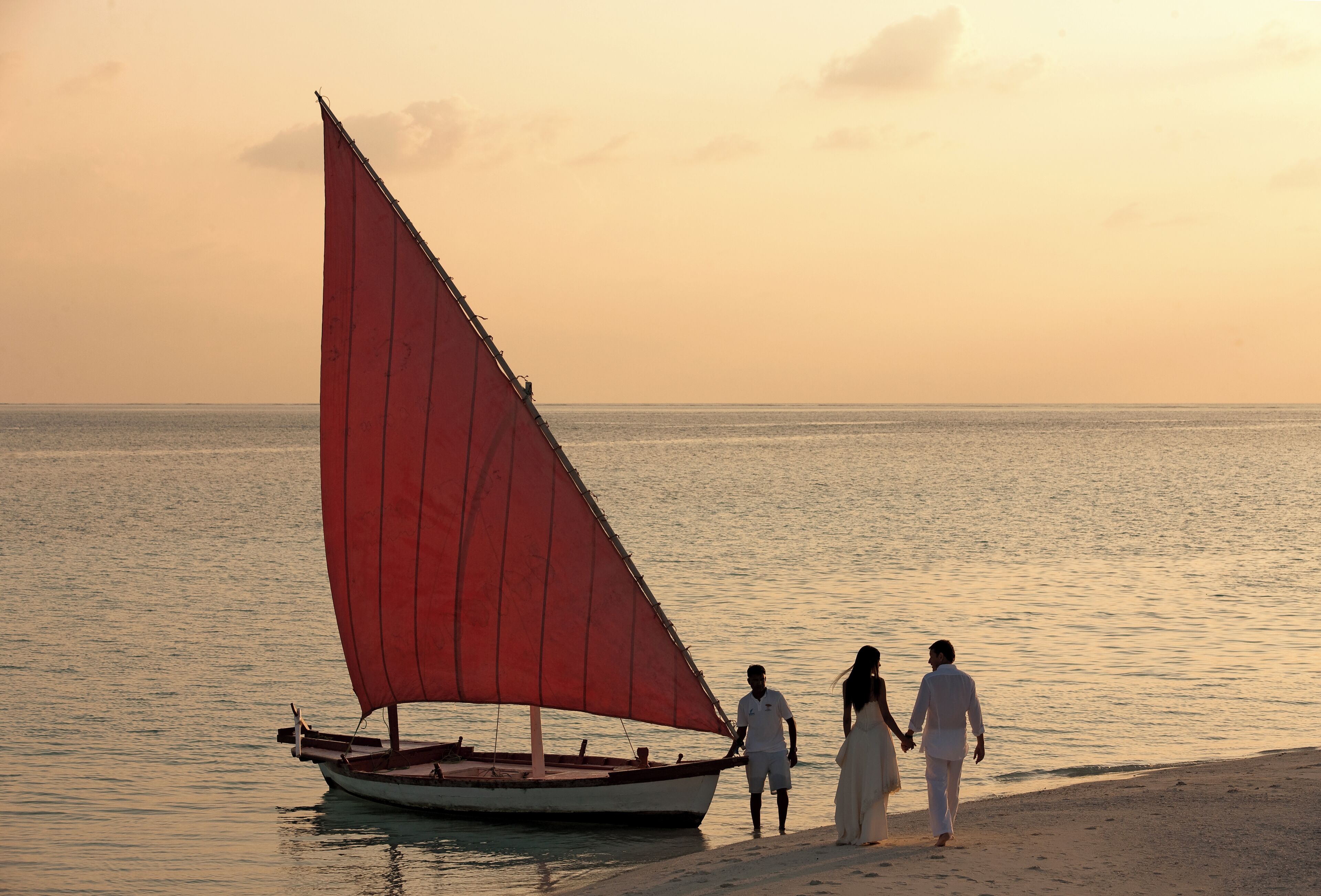 A boat with a red sail on calm water, a couple walking on a sandy shore, and a sunset in the background.