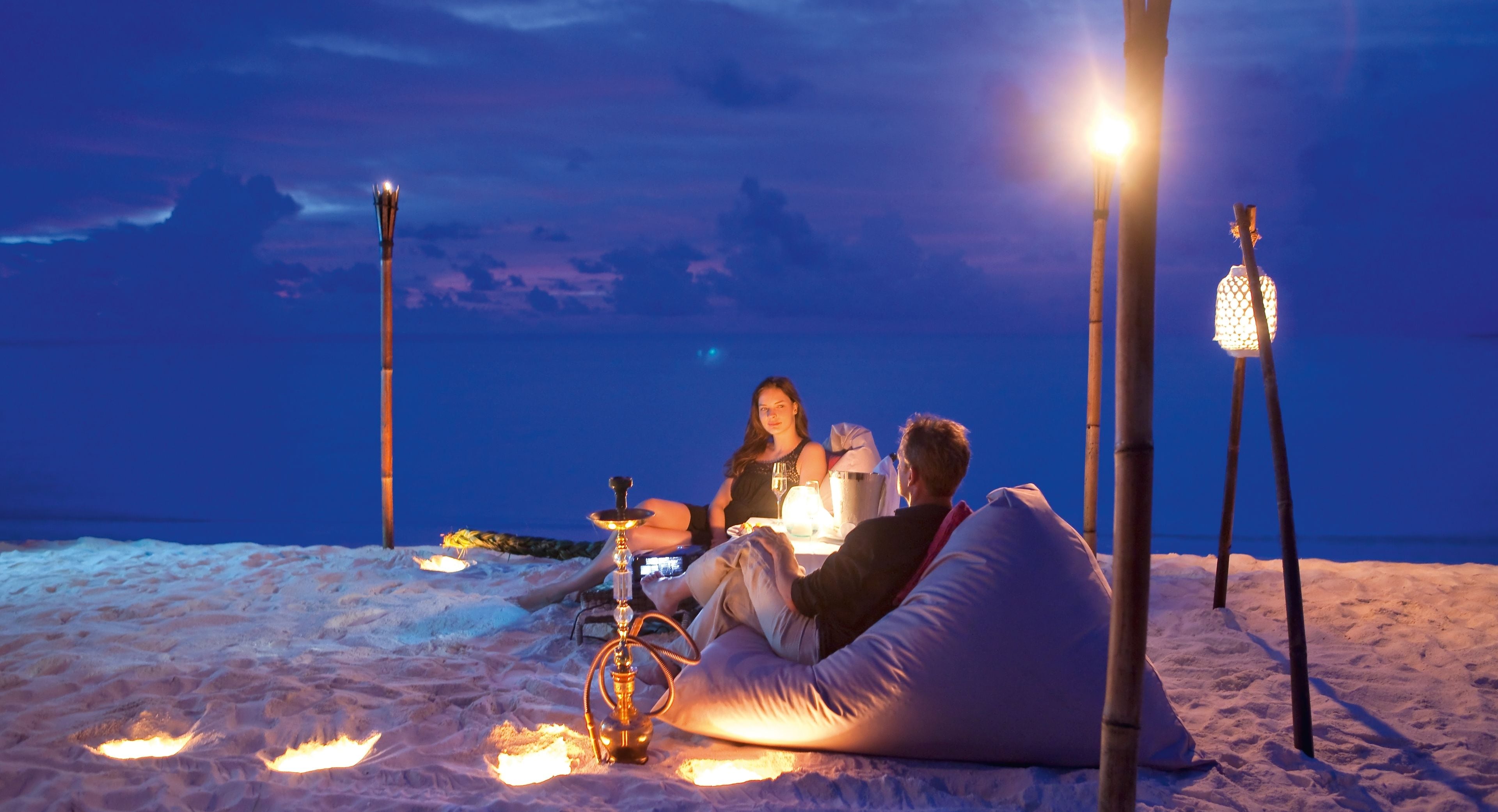 A beach scene with people relaxing on a bean bag, a hookah, and a lamp post.