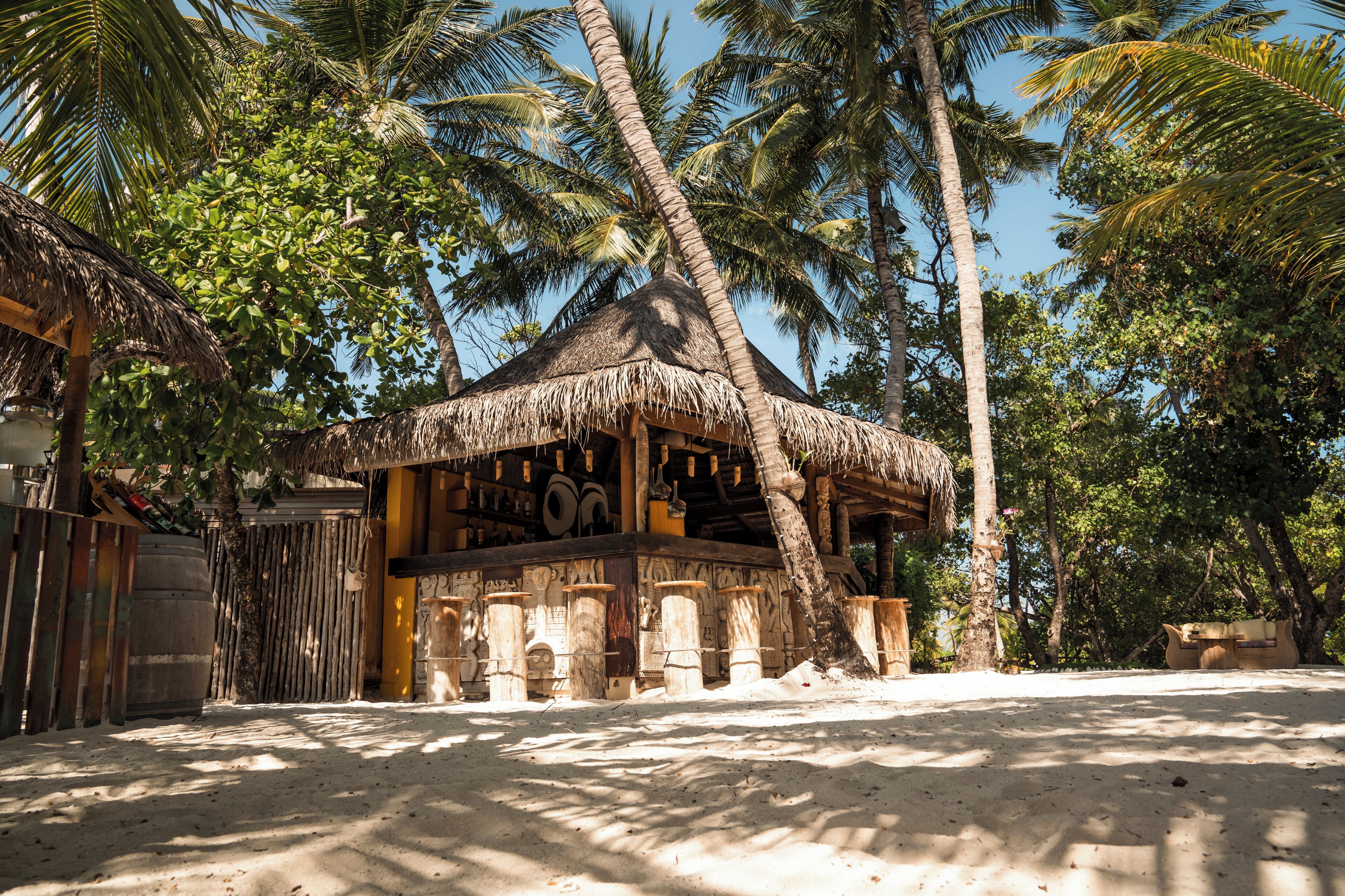 A thatched-roof structure with a wooden porch, surrounded by palm trees and a sandy ground.