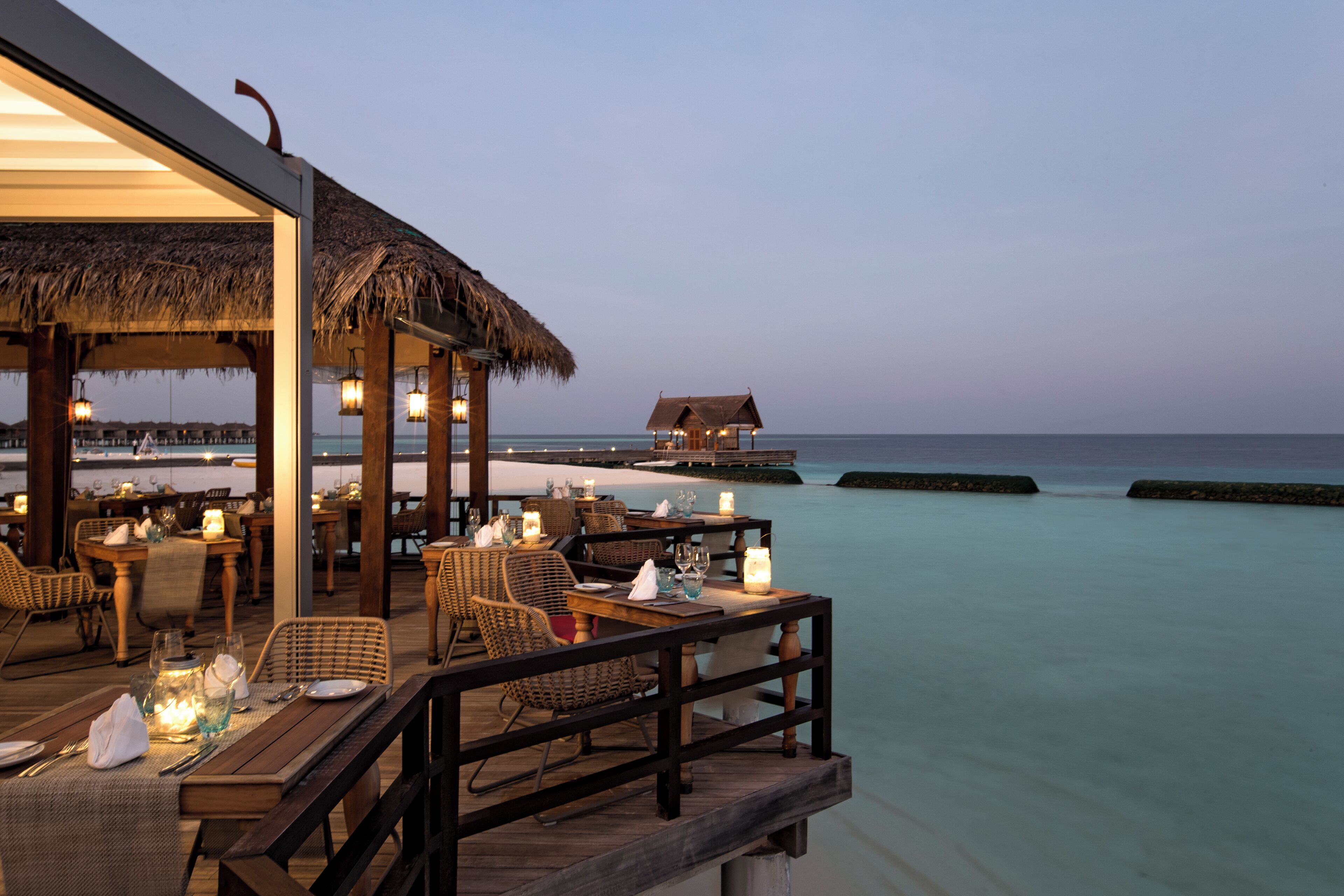 An outdoor dining area with tables and chairs, a thatched roof, and a view of the ocean.