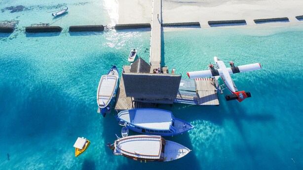 An aerial view of a floating structure with multiple boats docked, a jet bridge, and a sandy beach in the background. - Constance Moofushi All Inclusive (Moofushi Island)