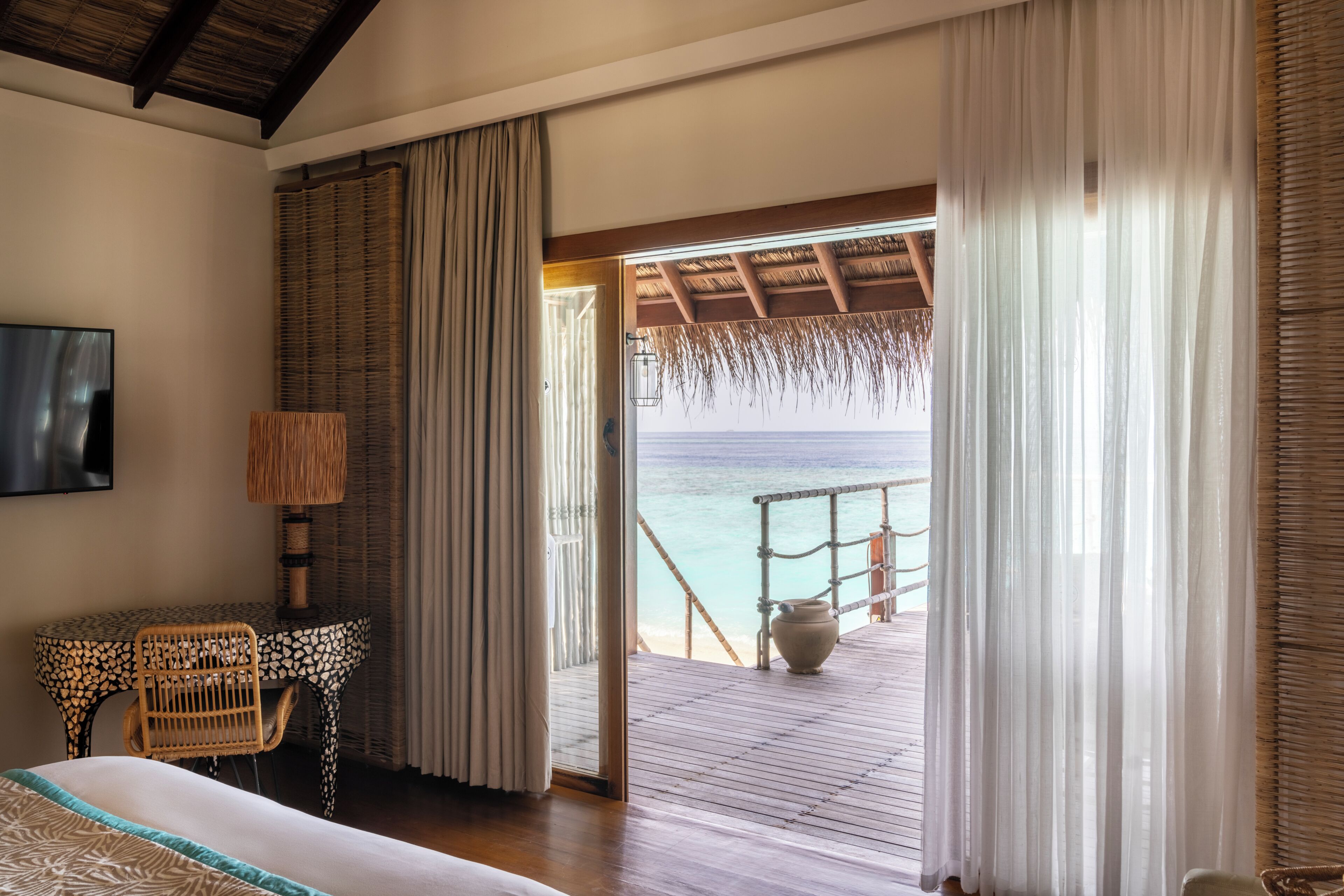Una habitación con cama, televisor, mesa y sillas, vista a la playa y terraza de madera.