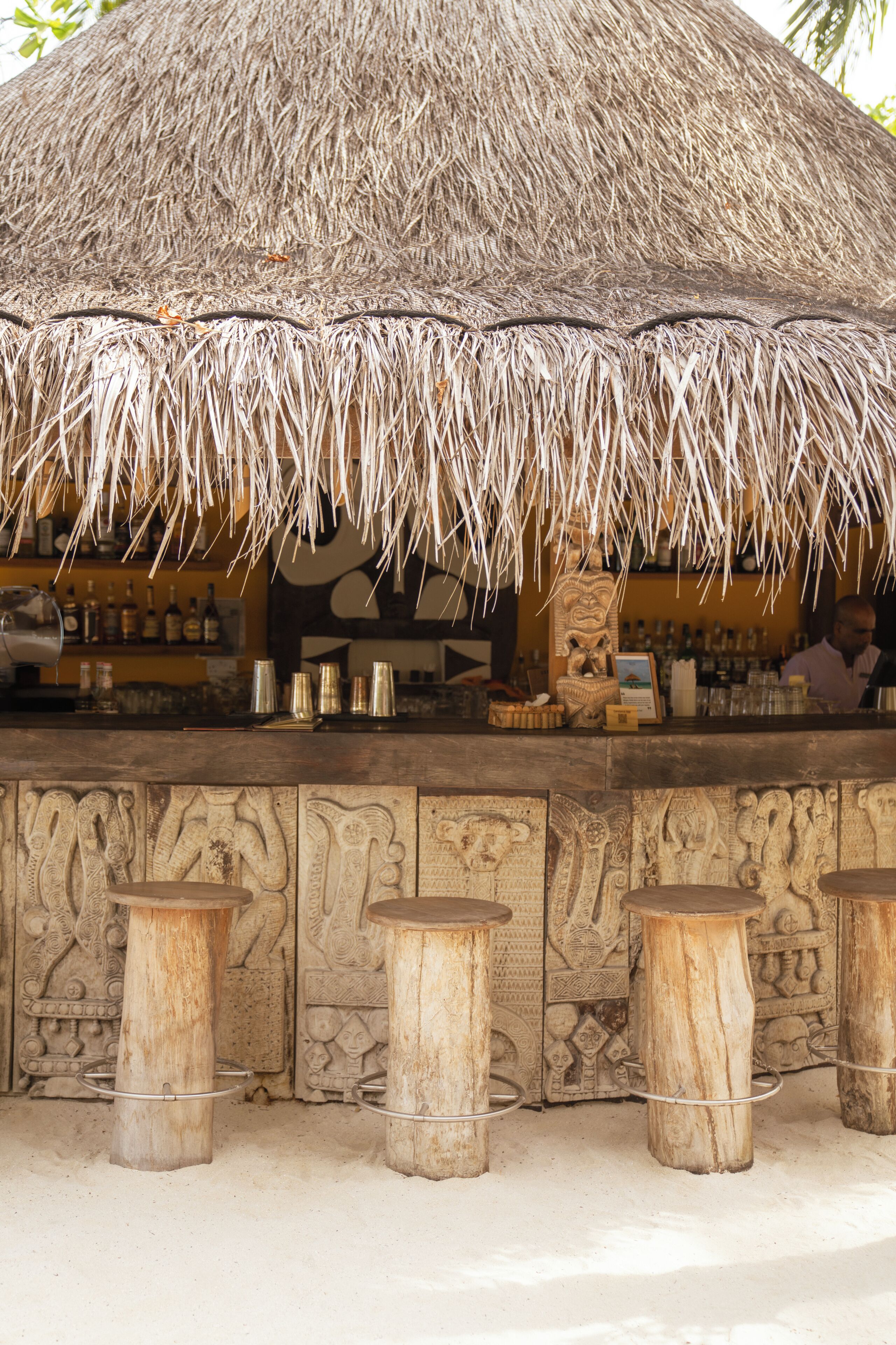 A bar area with a thatched roof, wooden stools, and a counter with various items.