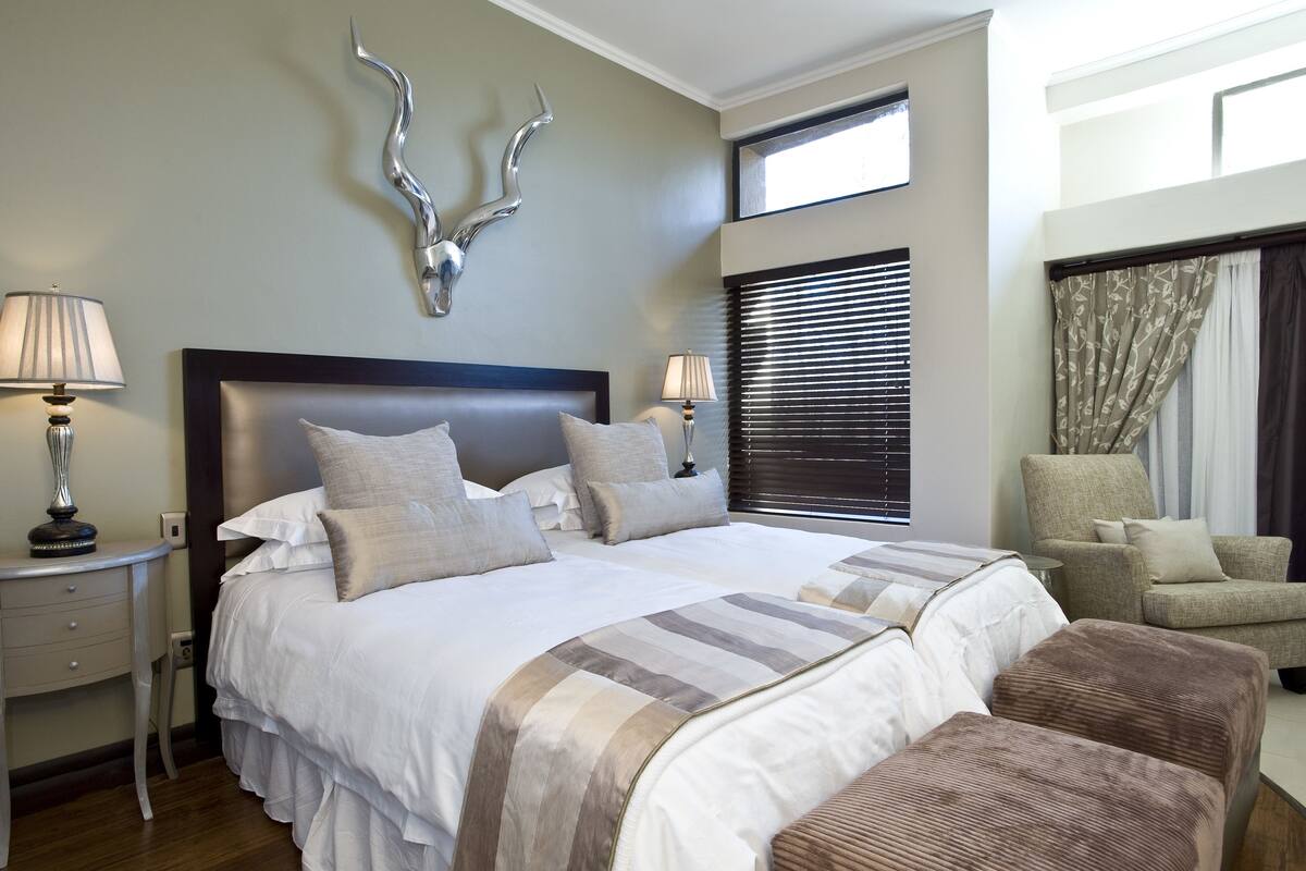A double bed with a headboard, two bedside tables with lamps, a chair, and a window with blinds.