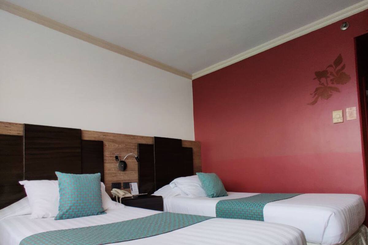 A hotel room with two beds, each with white bedding and teal accents. The room features a wooden headboard, a red accent wall, and a small bedside table with a lamp.