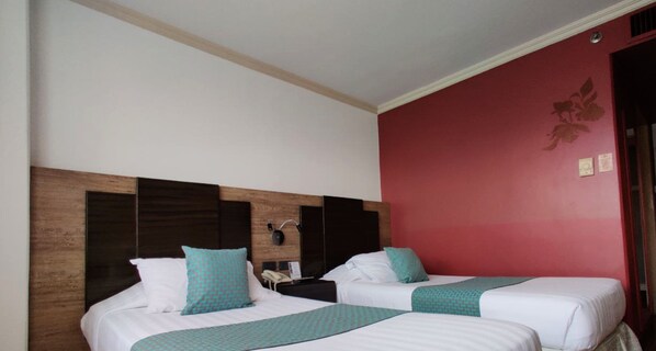 A hotel room with two beds, each with white bedding and teal accents. The room features a wooden headboard, a red accent wall, and a small bedside table with a lamp.