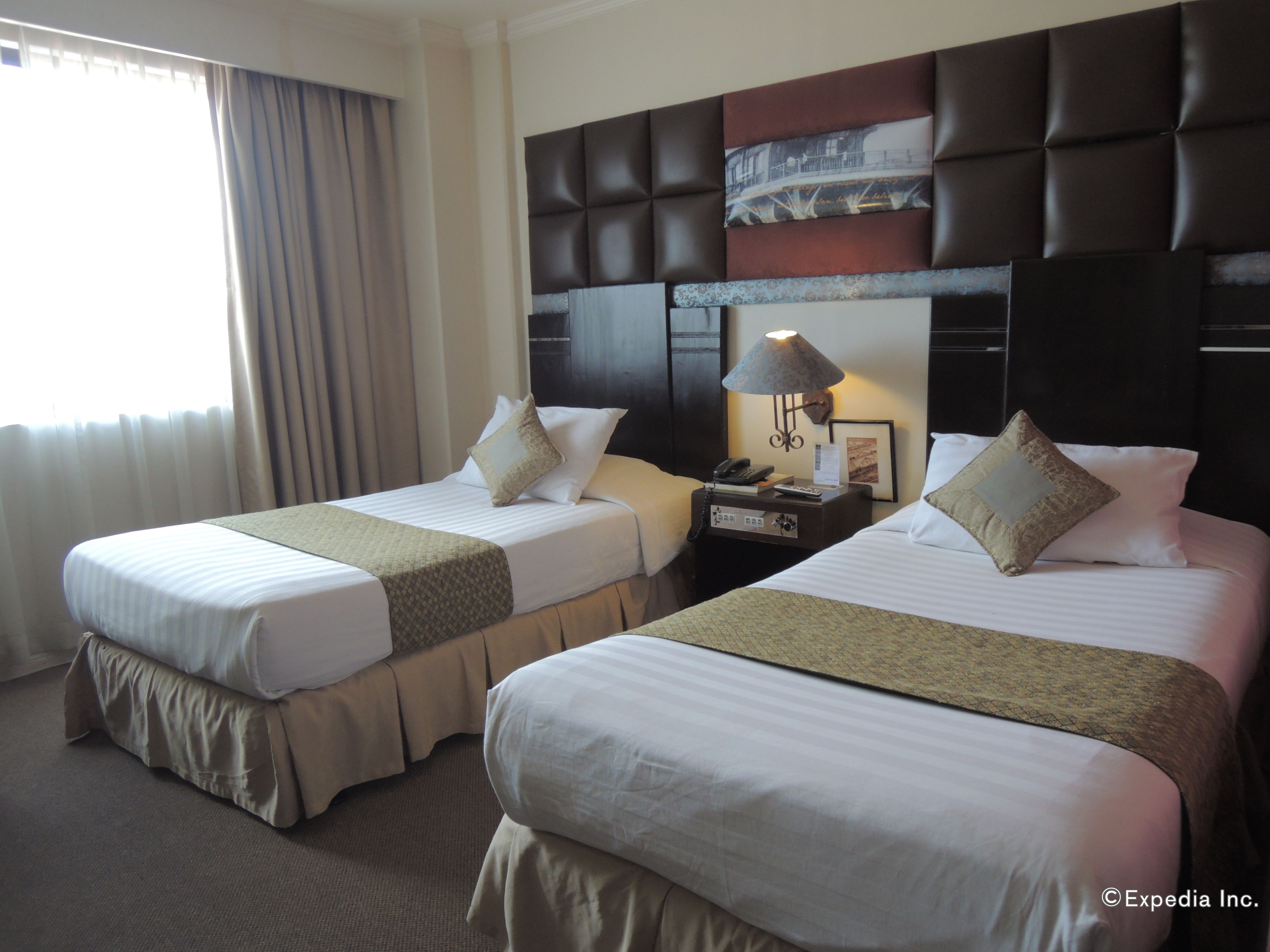 A hotel room with two beds, a nightstand with a lamp, and a headboard with a cityscape picture.
