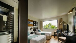 Habitación de hotel con dos camas, un escritorio con silla, televisión y ventana con vistas a zonas verdes.