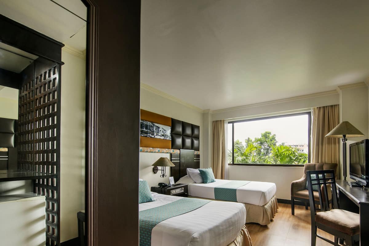 A hotel room with two beds, a desk with a chair, a television, and a window with a view of greenery.