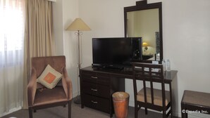 Una habitación de hotel con un sillón marrón, un escritorio con televisor, una lámpara y un cubo de basura.