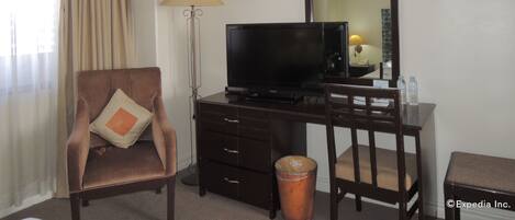 A hotel room with a brown armchair, a desk with a television, a lamp, and a trash can.