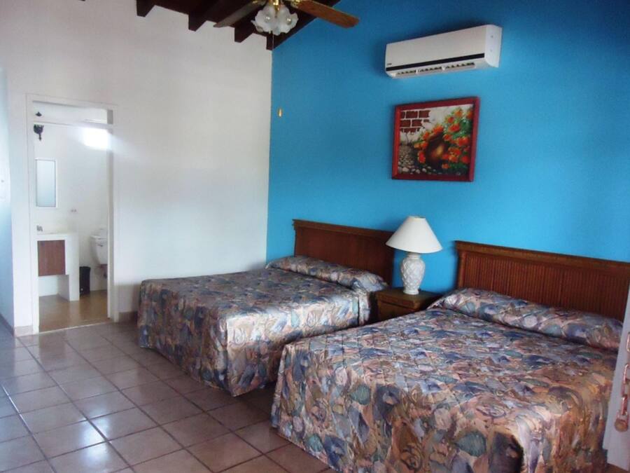 Quarto com duas camas, uma mesa de cabeceira com abajur, ventilador de teto, ar condicionado e um banheiro visível por uma porta aberta.
