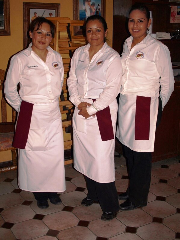 Three individuals in white chef uniforms with aprons standing in a room with tiled flooring and wooden furniture.