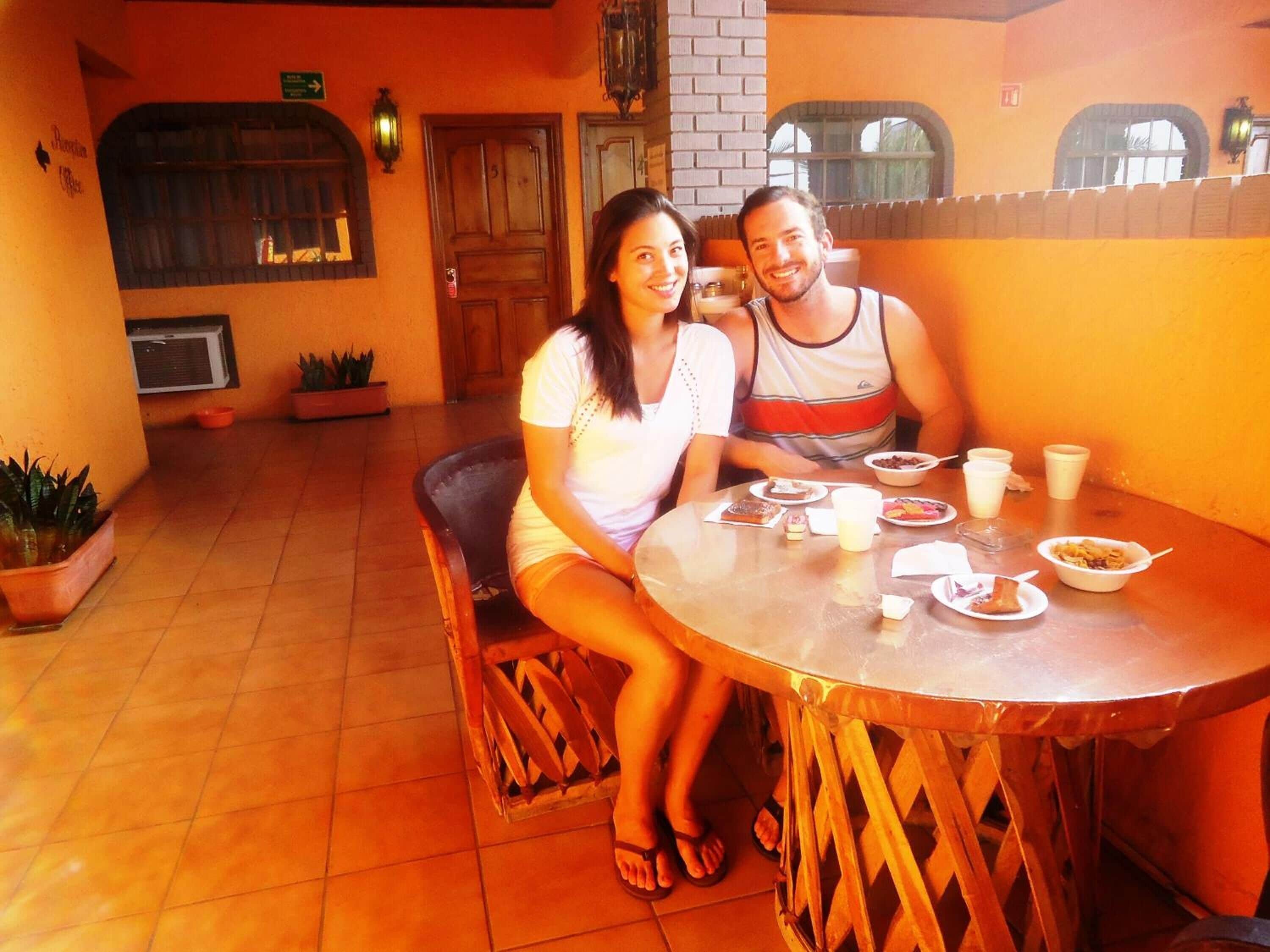 Una pareja sentada en una mesa redonda con varios platos y bebidas, en una habitación con piso de baldosas, plantas en macetas y una pared de ladrillo.