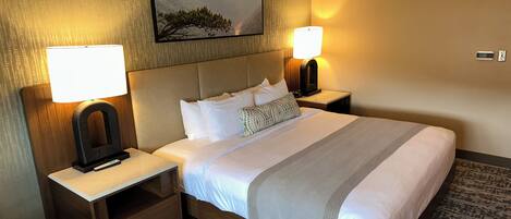 A hotel room with a large bed, two bedside tables, a nightstand, a lamp, a painting, and a fire alarm.