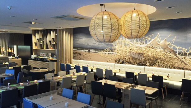 A modern restaurant with a beach mural, blue chairs, and hanging light fixtures. - Ostend Hotel (Ostend)