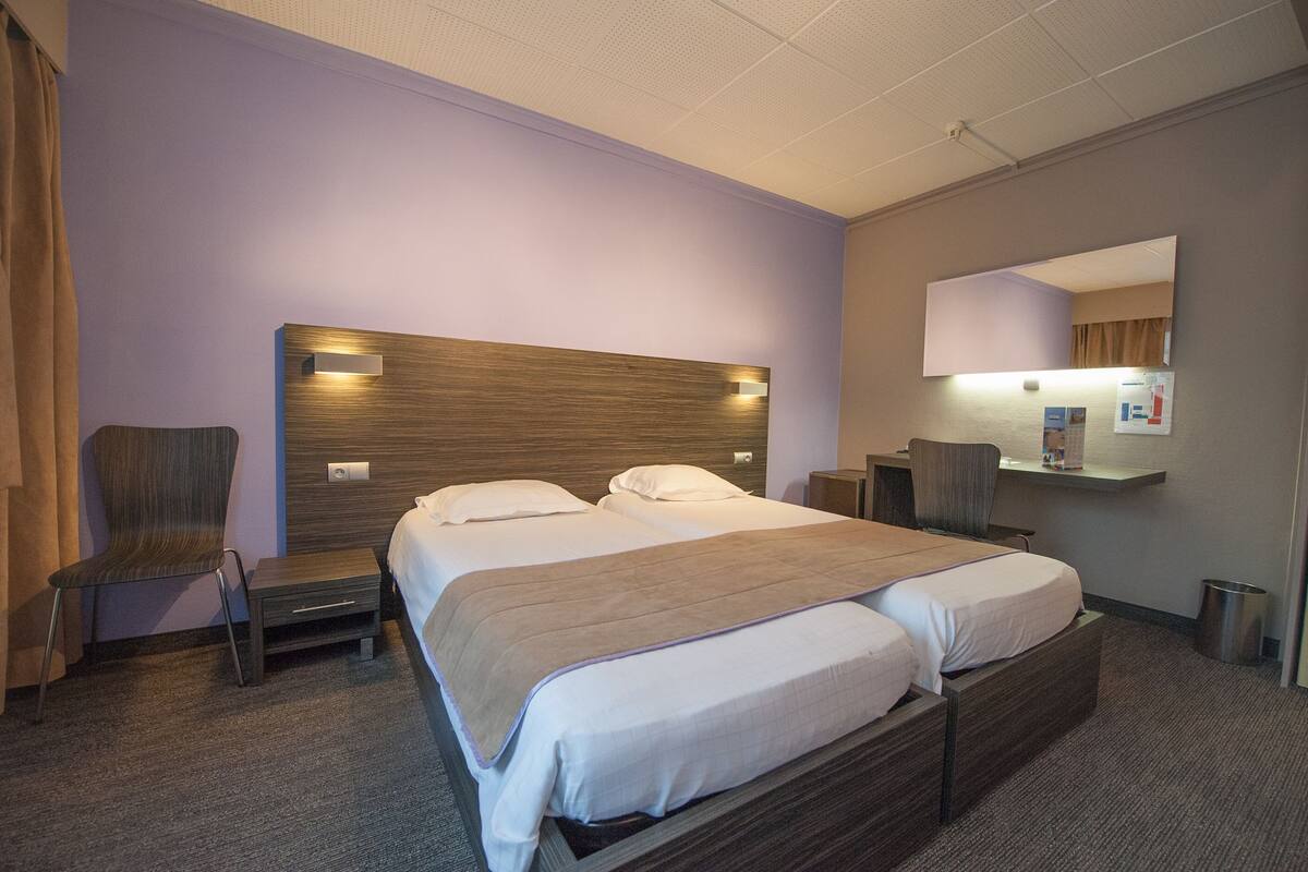 A hotel room with a large bed, two bedside tables, a chair, a small table, a mirror, and a wall-mounted shelf.