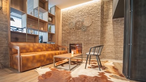 A cozy living room with a leather sofa, a coffee table, and a chair. There is a fireplace with a fire, a stone wall with decorative gears, and a glass door. - Ostend Hotel (Ostend)