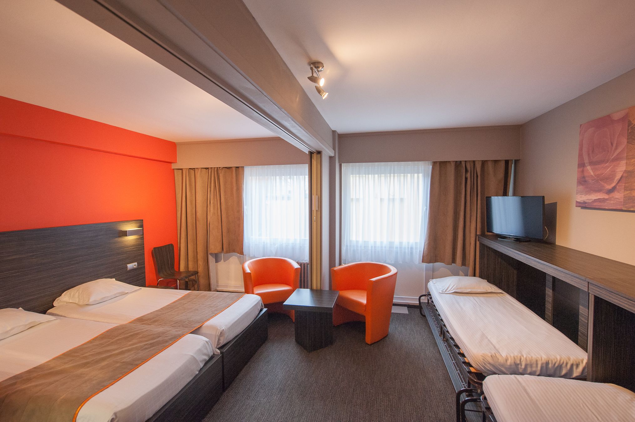 Ein Hotelzimmer mit zwei Betten, einem kleinen Tisch, zwei orangefarbenen Sesseln, einem Fernseher und einem Fenster mit Vorhängen.