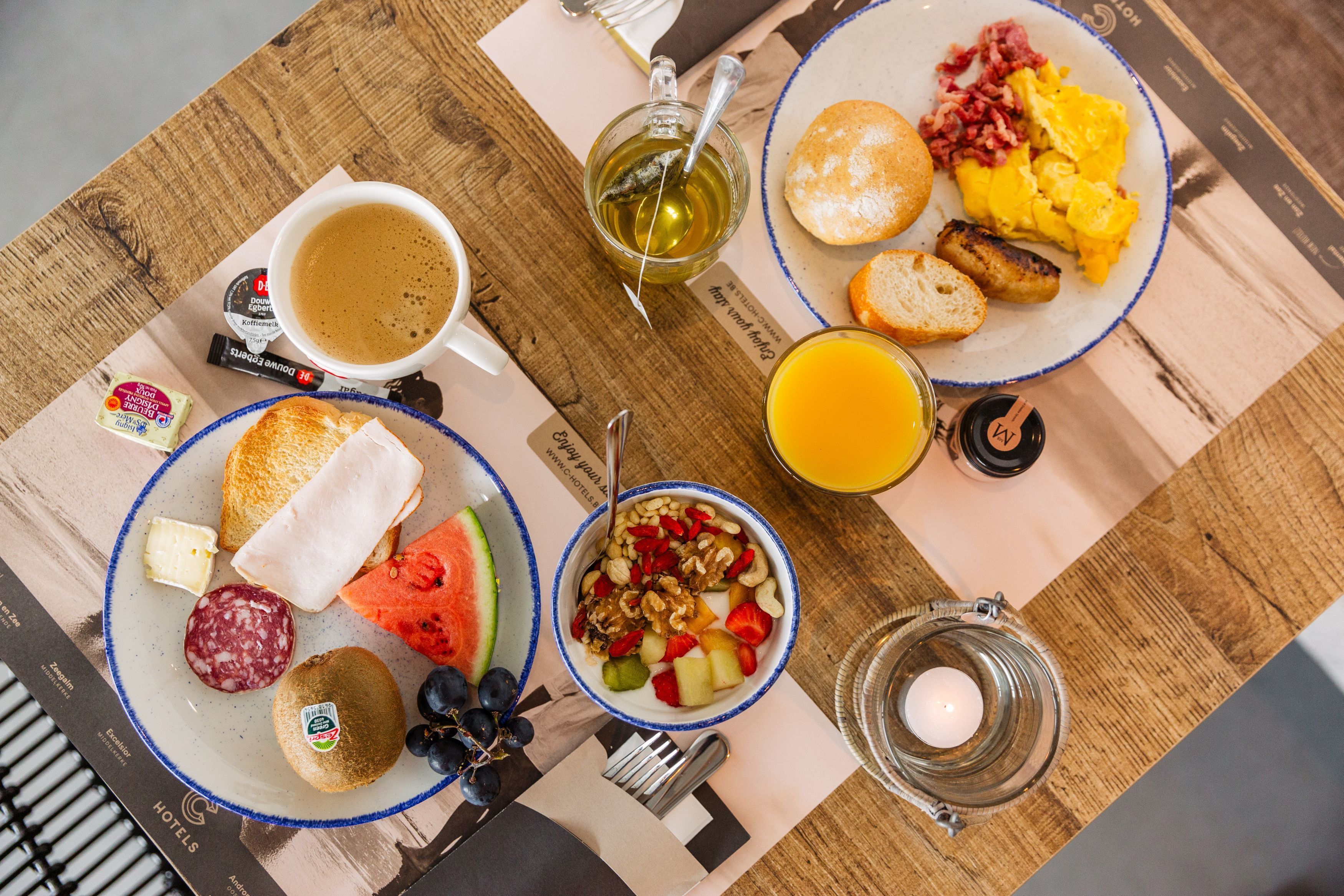 A breakfast table with plates of toast, eggs, bacon, and fruit. There is a cup of coffee and a glass of orange juice.