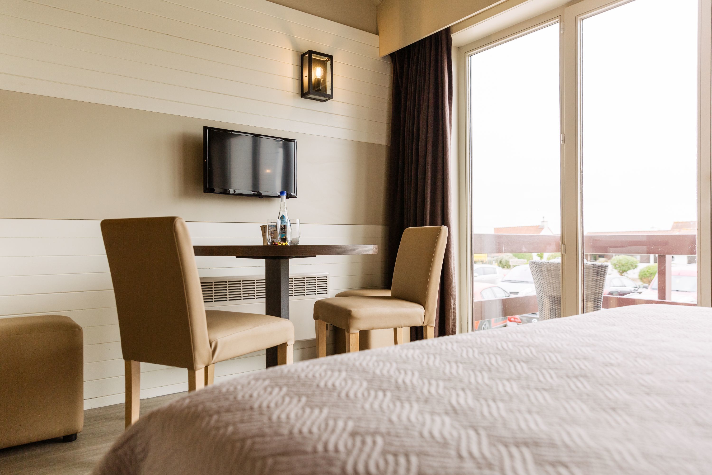 Une chambre d’hôtel avec un lit, deux chaises, une table avec une bouteille et des verres, un téléviseur fixé au mur et une grande fenêtre donnant sur un paysage.